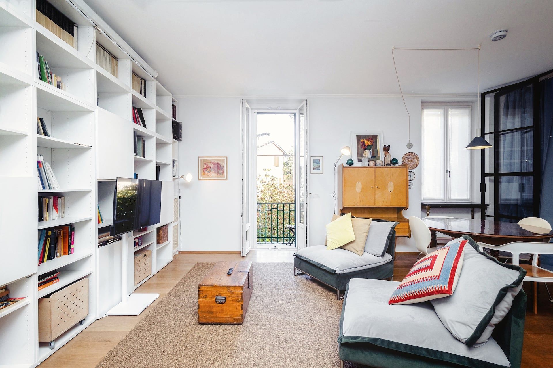Salotto luminoso e accogliente con libreria su misura, Smart TV con servizi streaming, due poltrone verdi, scrivania per lavorare, tavolo da pranzo e cucina annessa, il tutto su elegante parquet in legno naturale.