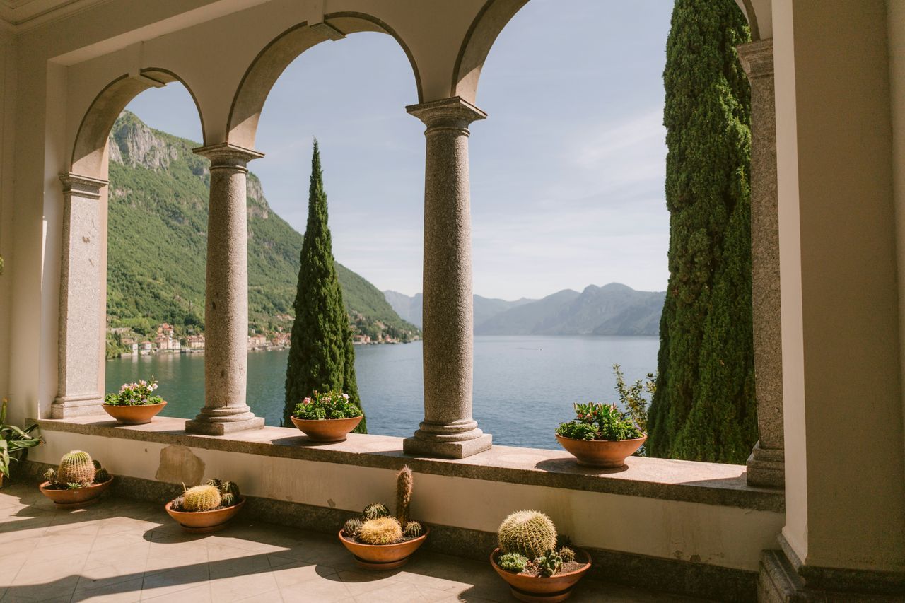 Loggia affacciata sul Lago di Como, tra colonne, vasi di fiori e cactus, con le colline e un borgo sull'acqua sullo sfondo.